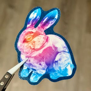 May include: A colorful bunny sticker with a gradient of pink, orange, and blue hues. The sticker has a dark blue glittery outline and is being held by a pair of tweezers. The bunny is a whimsical design.