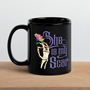 May include: Black ceramic mug featuring a skeletal hand holding a flower with pink and green petals. The mug has the text "She is my Scar" in purple. The mug is on a wooden surface.