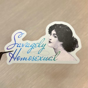 May include: A white sticker with a black and white image of a woman with dark hair. The text "Savagely Homosexual" is written in a rainbow gradient.