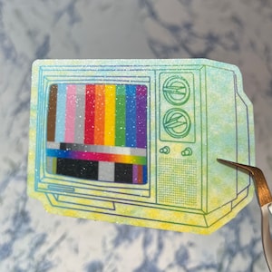 May include: A vintage-style television with a glittery blue and yellow background. The screen shows a colorful test pattern.