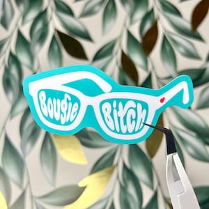 May include: A turquoise sticker in the shape of sunglasses with the words "Bougie Bitch" in white script. A small red heart is on the right side of the glasses. The sticker is held by tweezers against a background of green leaves.