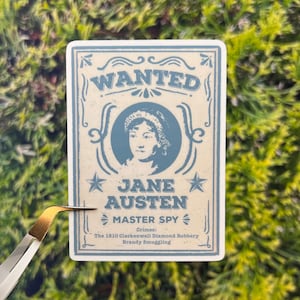 May include: A vintage-style wanted poster with a blue and white color scheme. The poster features a portrait of Jane Austen, with the text "WANTED", "JANE AUSTEN", "MASTER SPY", "Crimes: The 1810 Clerkenwell Diamond Robbery, Brandy Smuggling".