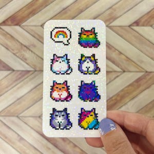 May include: A sticker sheet with eight pixelated cat designs in different colours. One design shows a rainbow over the letter "Q". The other designs feature cats in rainbow, blue, pink, orange, purple, and grey colour schemes. The sticker sheet has a glittery finish.