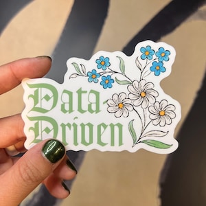 May include: A white sticker with green text that reads "Data Driven" in a gothic font. The text is surrounded by a floral design with blue and white flowers.