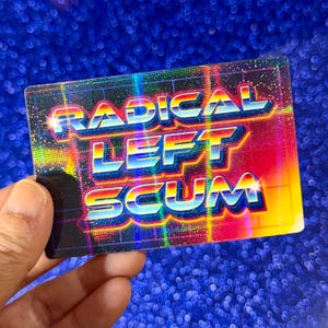 May include: A rectangular sticker with the words "RADICAL LEFT SCUM" in a retro, neon-style font. The text is in shades of blue, pink, and yellow, with a grid pattern in the background. The sticker has a holographic, iridescent finish.
