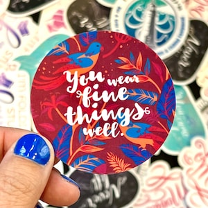 May include: A round sticker with a red, blue, and orange floral design. The sticker features the text "You wear fine things well."