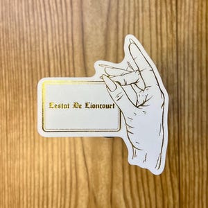 May include: A white card with gold trim and the text "Lestat De Lioncourt" in gold lettering. A hand with long fingers is holding the card.