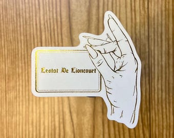 May include: A white card with gold trim and the text "Lestat De Lioncourt" in gold lettering. A hand with long fingers is holding the card.
