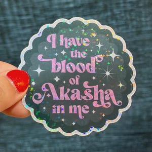 May include: A scalloped circular sticker with a black background and holographic glitter accents. The text reads I have the blood of Akasha in me in a stylized pink and purple gradient font, surrounded by small white four-pointed star icons.