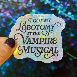 May include: A die-cut sticker with a holographic finish featuring a star pattern and the black text I GOT MY LOBOTOMY AT THE VAMPIRE MUSICAL. The sticker has an irregular, wavy edge and a reflective surface that catches light in various colors.