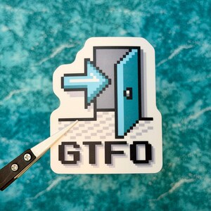 May include: A white sticker with a pixelated design featuring a blue arrow pointing towards a blue door. The letters "GTFO" are printed in a pixelated font below the door graphic. A pair of tweezers is holding the sticker.
