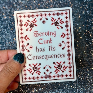 Peut inclure: Un autocollant rectangulaire blanc avec une bordure et des motifs floraux de style point de croix rouge foncé. Le texte "Serving Cunt has its Consequences" est imprimé au centre dans une police rouge foncé imitant la broderie traditionnelle.