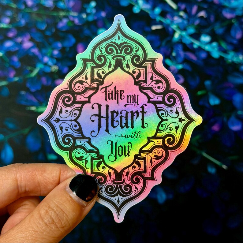 May include: A holographic sticker with a decorative border and the text "Take my Heart with You." The sticker has a rainbow gradient of colors and a black border. The background is a blurred image of blue flowers.