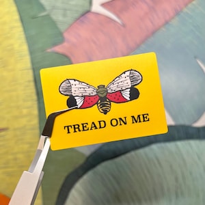 May include: A yellow rectangle sticker with a moth illustration and the text "TREAD ON ME" in black. The moth has white, red, and black wings. The sticker is held by silver tweezers.