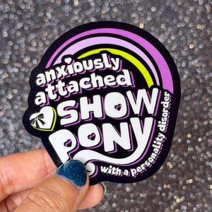 May include: A die-cut sticker with a black background featuring a rainbow arch in purple, pink, and yellow. The white text reads "anxiously attached SHOW PONY with a personality disorder." A white heart icon with a black bow is on the left side.