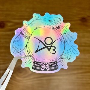 May include: A sticker with a holographic finish featuring a hand holding a crystal ball with a tie-dye pattern and the logo "Y3" inside. The hand is adorned with bracelets and a star tattoo.