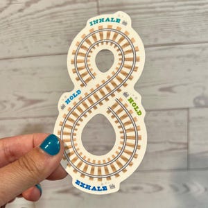 May include: A cream-coloured sticker in the shape of an infinity symbol, with a railway track design. The words "INHALE," "HOLD," and "EXHALE" are printed on the sticker in teal and green. The sticker is held by a hand with teal nail polish.