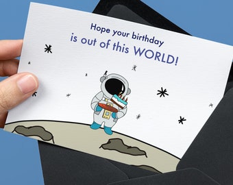 May include: A white card with a cartoon astronaut holding a birthday cake. The card says "Hope your birthday is out of this WORLD!" and has a black envelope.