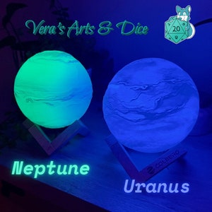 Może przedstawiać: Dwie podświetlane lampy w kształcie księżyca, jedna zielona, a druga niebieska, z drewnianą podstawą. Zielona lampa jest oznaczona jako "Neptun", a niebieska jako "Uran".