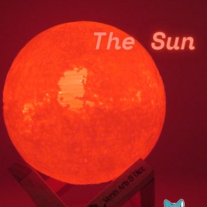 Op de afbeelding: Een rode maanlamp met een getextureerd oppervlak, verlicht en op een houten standaard. De lamp is gelabeld "The Sun" en is een product van Vera's Arts & Dice.