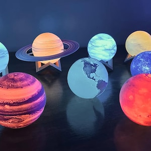 Op de afbeelding: Een verzameling verlichte decoratieve lampen in de vorm van planeten. De lampen zijn in verschillende kleuren, waaronder blauw, oranje en rood, en worden op kleine houten standaards gepresenteerd. Eén lamp heeft een ring, die op Saturnus lijkt.