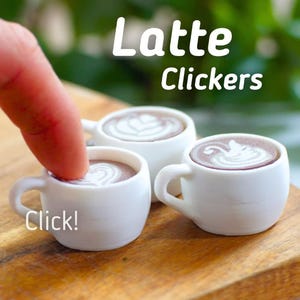 Könnte beinhalten: Drei Miniatur-Kaffeetassen aus weißer Keramik mit Latte-Art-Designs. Die Tassen stehen auf einer Holzoberfläche. Der Text "Latte Clickers" befindet sich über den Tassen. Der Text "Click!" befindet sich unter den Tassen.