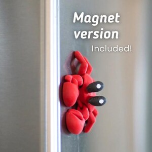 Puede incluir: Un im&aacute;n rojo con forma de cangrejo con ojos negros y blancos est&aacute; adherido a un refrigerador plateado. El texto "Magnet version Included!" se muestra en blanco.
