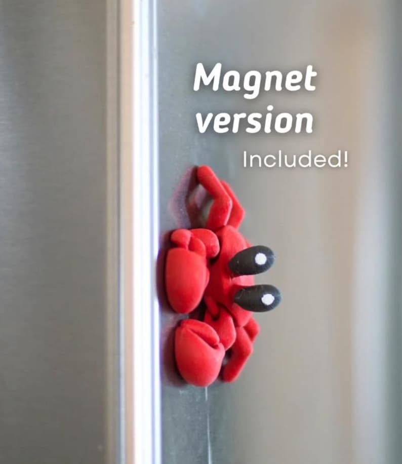 Puede incluir: Un im&aacute;n rojo con forma de cangrejo con ojos negros y blancos est&aacute; adherido a un refrigerador plateado. El texto "Magnet version Included!" se muestra en blanco.