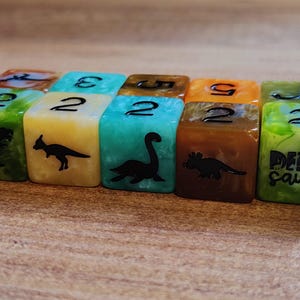 Puede incluir: Un conjunto de diez dados coloridos con ilustraciones de dinosaurios. Los dados están numerados del 2 al 12. Los dados están hechos de un material translúcido con remolinos de verde, azul, amarillo y naranja. Los dados están etiquetados como "Dino Saurs".