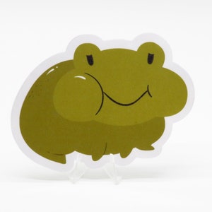 Puede incluir: Una pegatina de rana verde con una cara sonriente y los ojos cerrados. La rana tiene una superficie texturizada y está delineada en blanco.