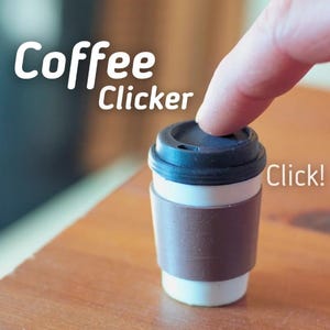 May include: A small coffee cup with a brown sleeve and a black lid. The words "Coffee Clicker" are printed on the image, with the word "Click!" next to the cup. A finger is pressing down on the lid.