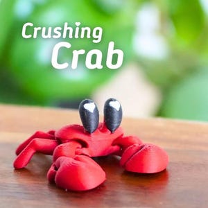 Puede incluir: Figura de cangrejo rojo impresa en 3D con ojos negros y blancos. El cangrejo tiene grandes pinzas y est&aacute; posado sobre una superficie de madera. El texto "Crushing Crab" est&aacute; en letras blancas encima del cangrejo.