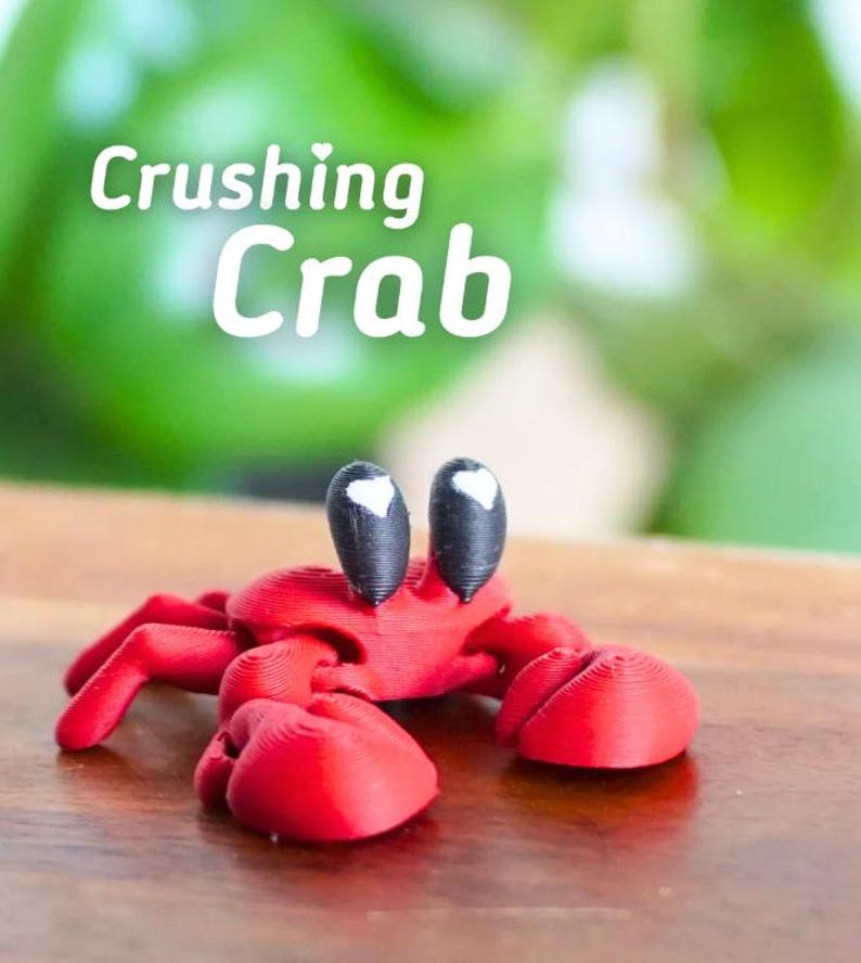 Puede incluir: Figura de cangrejo rojo impresa en 3D con ojos negros y blancos. El cangrejo tiene grandes pinzas y est&aacute; posado sobre una superficie de madera. El texto "Crushing Crab" est&aacute; en letras blancas encima del cangrejo.
