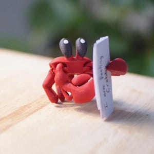 Puede incluir: Figura de cangrejo rojo con ojos negros, sosteniendo un letrero blanco que dice "I am feeling crabby". El cangrejo sujeta el letrero con sus pinzas. La figura est&aacute; sobre una superficie de madera clara.