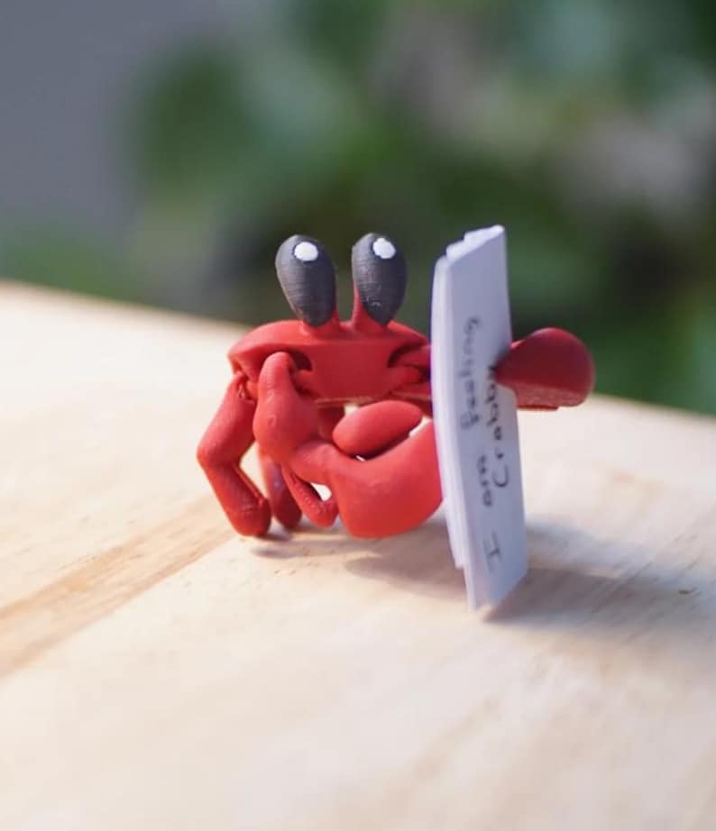 Puede incluir: Figura de cangrejo rojo con ojos negros, sosteniendo un letrero blanco que dice "I am feeling crabby". El cangrejo sujeta el letrero con sus pinzas. La figura est&aacute; sobre una superficie de madera clara.