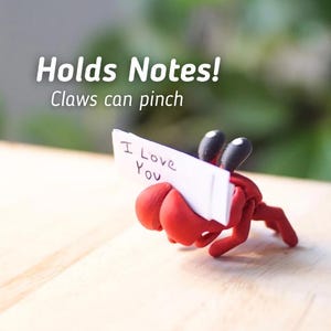 Puede incluir: Una figura de cangrejo de arcilla roja que sostiene una peque&ntilde;a tarjeta blanca que dice "I Love You". El cangrejo tiene ojos negros y est&aacute; colocado sobre una superficie de madera. El texto encima del cangrejo dice "Holds Notes! Claws can pinch."