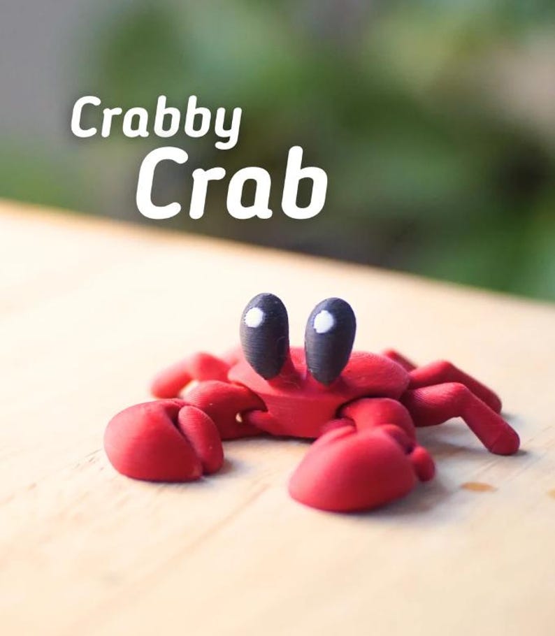 Puede incluir: Una figura de cangrejo rojo hecha a mano con grandes ojos negros y reflejos blancos. El cangrejo tiene grandes pinzas y est&aacute; sobre una superficie de madera. El texto "Crabby Crab" est&aacute; escrito en blanco encima del cangrejo.