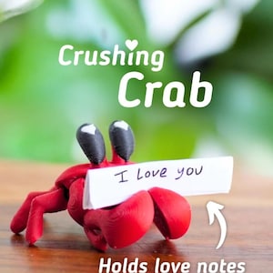 Puede incluir: Una figura de cangrejo rojo con ojos negros, sosteniendo un letrero blanco que dice "I Love you". El cangrejo sostiene el letrero con sus pinzas. Tambi&eacute;n se ve el texto "Crushing Crab" y "Holds love notes".