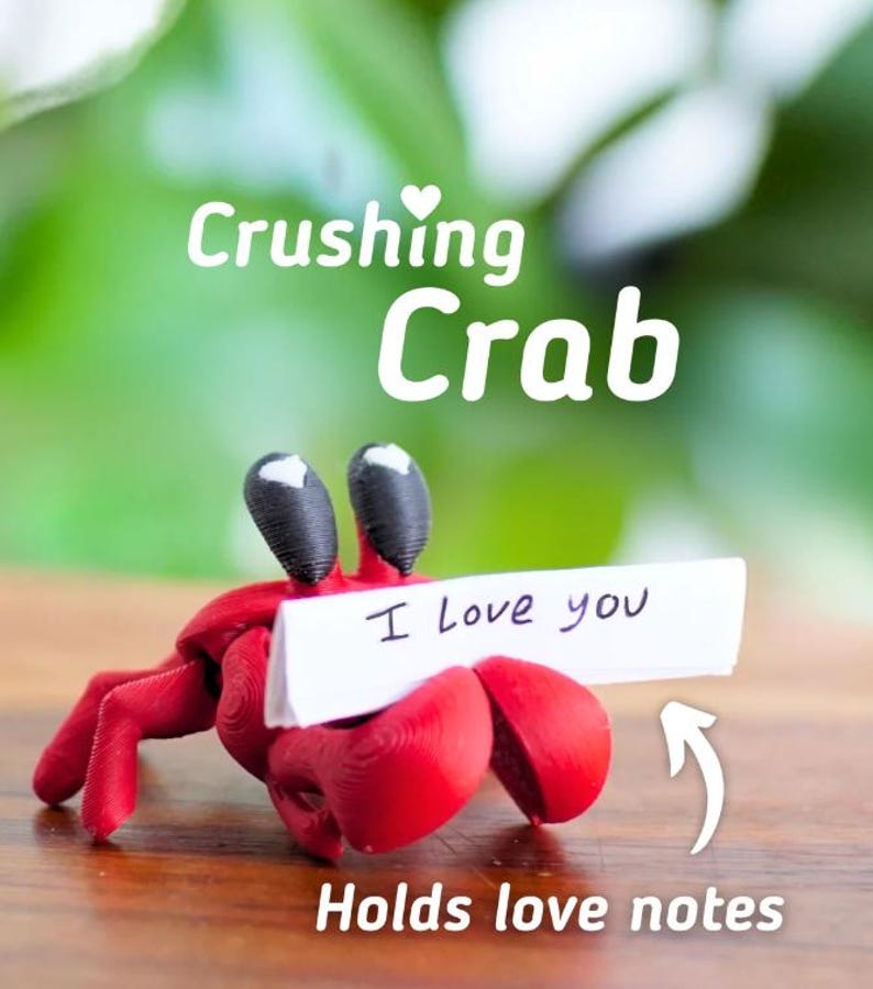 Puede incluir: Una figura de cangrejo rojo con ojos negros, sosteniendo un letrero blanco que dice "I Love you". El cangrejo sostiene el letrero con sus pinzas. Tambi&eacute;n se ve el texto "Crushing Crab" y "Holds love notes".