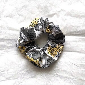 Könnte beinhalten: Ein floraler Haargummi mit einem Muster aus grauen und gelben Blumen auf weißem Hintergrund. Der Stoff ist gerafft und gebunden, wodurch eine kreisförmige Form entsteht. Das Haargummi ist für das Haarstyling konzipiert.