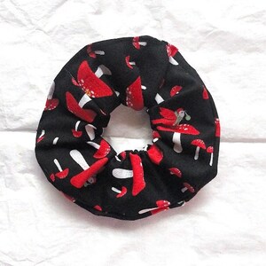 Könnte beinhalten: Schwarzes Haargummi mit einem sich wiederholenden Muster aus roten und weißen Pilzen. Das Haaraccessoire ist kreisförmig und gerafft. Die Pilze haben rote Kappen und weiße Stiele auf schwarzem Hintergrund.