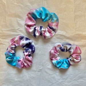 May include: Three tie-dye scrunchies in shades of blue, pink, and purple. The fabric is soft and gathered, creating a textured appearance. The scrunchies are arranged on a light-colored, textured surface.