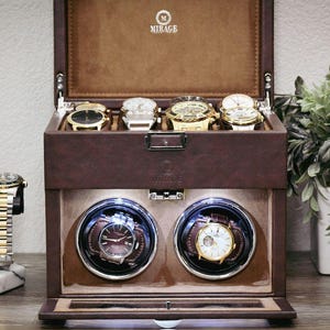 May include: A brown leather watch box with multiple compartments, holding several wristwatches. The box has a top section for storage and a lower section with watch winders. A wristwatch rests on a stand to the left. A plant in a white pot is on the right.