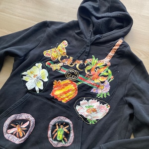 May include: Black hooded sweatshirt with a colorful floral and insect print. The print features butterflies, flowers, a snake, a bee, and a beetle. The print is surrounded by a Gucci logo.