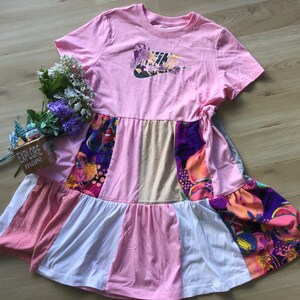 May include: A pink and white tiered dress with a Nike logo on the front. The dress has a patchwork design with different colored fabrics and patterns.