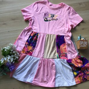 Może przedstawiać: R&oacute;żowo-biała sukienka w stylu patchwork z logo Nike na piersi. Sukienka ma kwiatowy wz&oacute;r na dole i jest wykonana z miękkiej, wygodnej tkaniny.