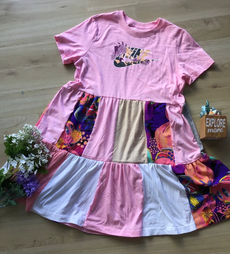 Może przedstawiać: R&oacute;żowo-biała sukienka w stylu patchwork z logo Nike na piersi. Sukienka ma kwiatowy wz&oacute;r na dole i jest wykonana z miękkiej, wygodnej tkaniny.