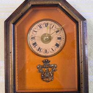May include: A vintage wall clock with a wooden frame and a light brown face. The clock features Roman numerals and decorative hands. A metal crest is below the clock face. The clock is approximately 20 cm tall.