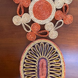 May include: Two woven trivets made of coiled paper or fiber. The top trivet is circular with a central orange disc surrounded by alternating beige and orange loops. The bottom trivet is oval with a central orange oval, yellow petals, and a purple border.