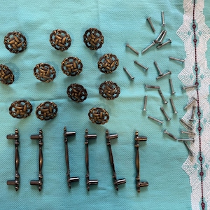 May include: A collection of antique bronze cabinet hardware, including decorative knobs and pulls, along with silver screws. The knobs feature an intricate woven design. The hardware is arranged on a light blue fabric.